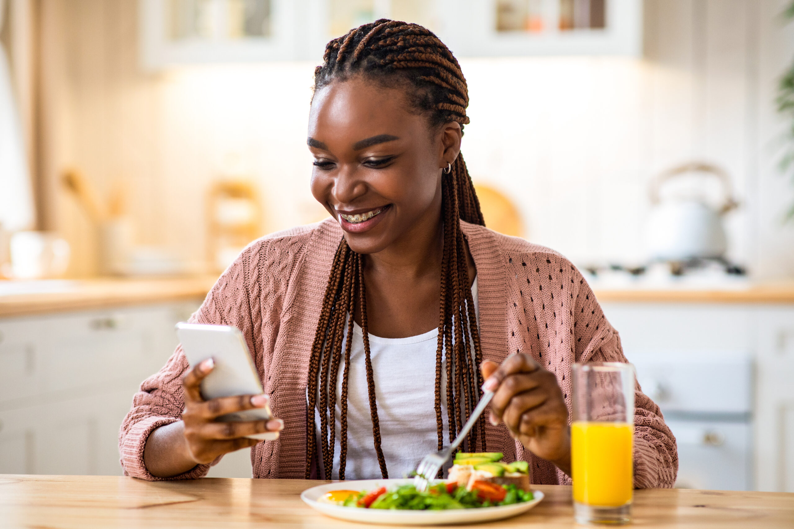 Smiling black woman using smartphone while having breakfast in kitchen at home